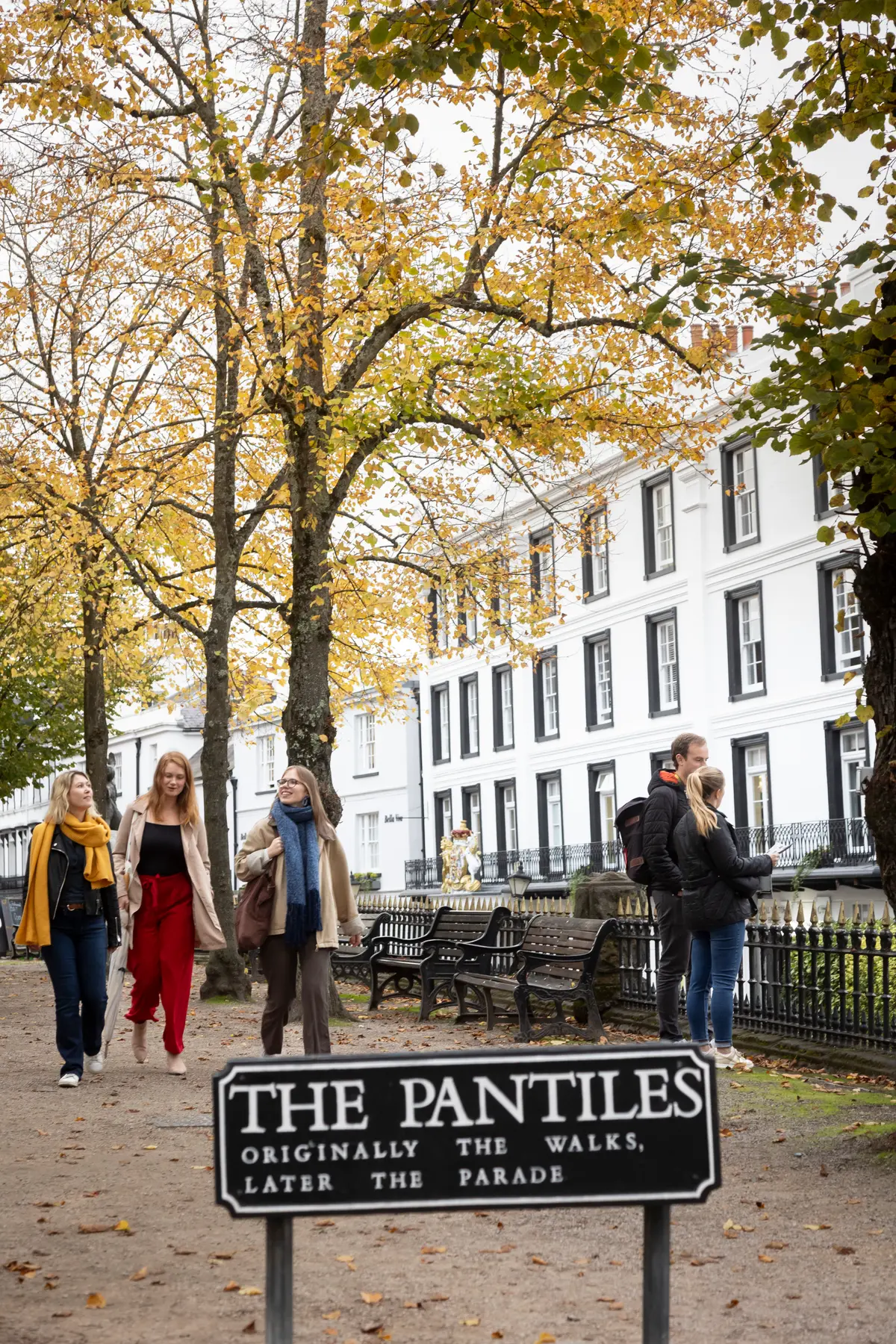 People walking along The Pantiles in Tunbridge Wells beneath autumnal trees, the white colonnade and the engraved Pantiles sign in view